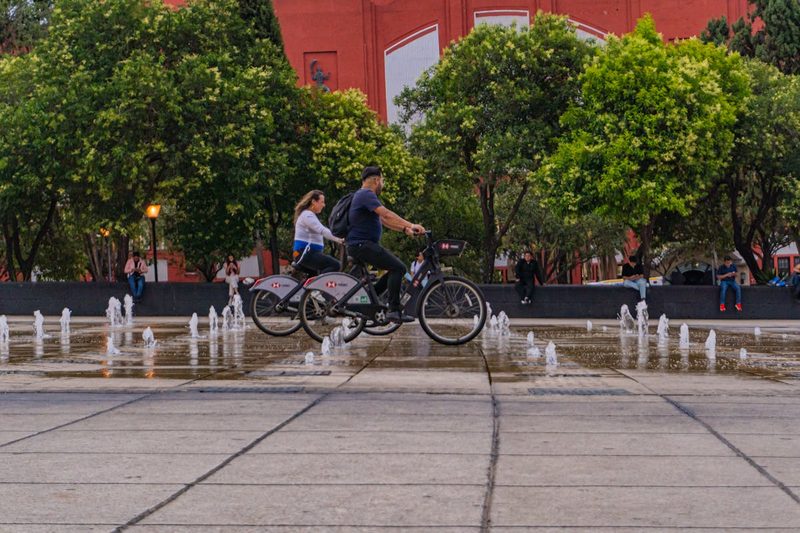 Two cyclists ride past fountains in a vibrant Mexico City park surrounded by lush greenery